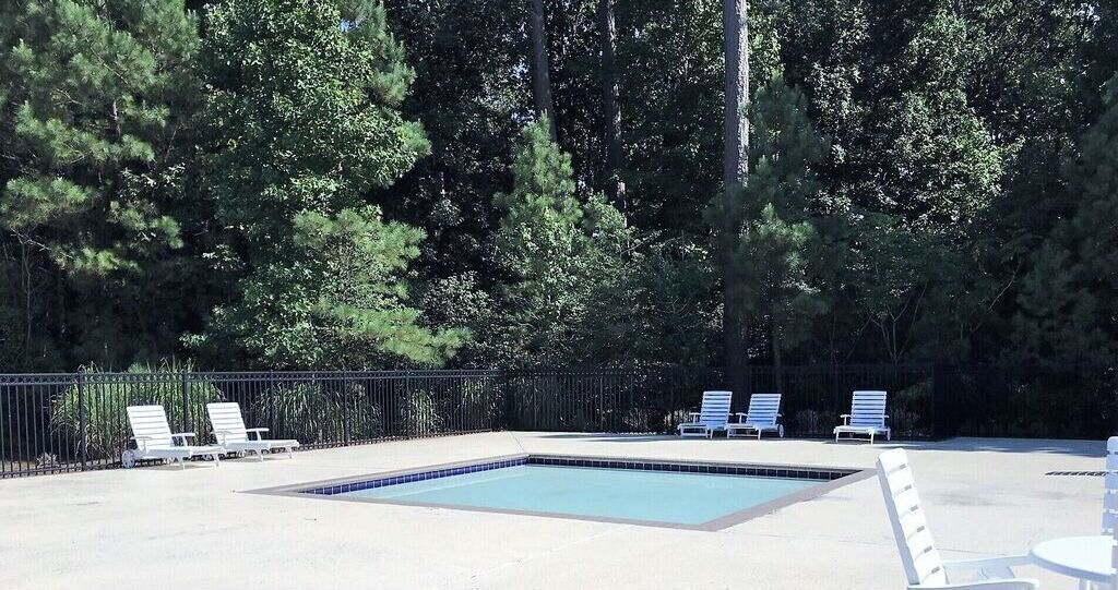 A rectangular swimming pool with a tiled edge, surrounded by a concrete deck. There are four white lounge chairs aligned along the pool's edge.
