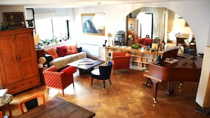 Ein gemütliches Wohnzimmer mit Holzboden, einer Couch, einem Couchtisch und einem Esstisch. Ein Bücherregal voller Bücher und ein Spiegel, der den Raum widerspiegelt.