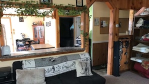 Un salon chaleureux avec un canapé noir, une table basse en bois et un coin cuisine visible à travers une porte ouverte.