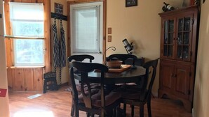 Un coin repas comprenant une table ronde, six chaises, une fenêtre avec des stores, un buffet en bois et un petit tabouret.