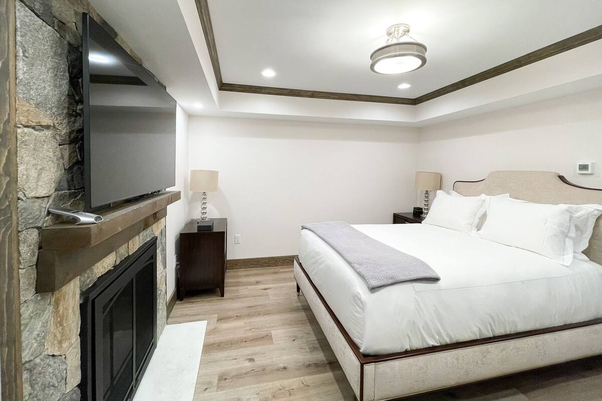 A bedroom with a bed, a TV mounted on a stone wall, a nightstand with a lamp, and a ceiling light.
