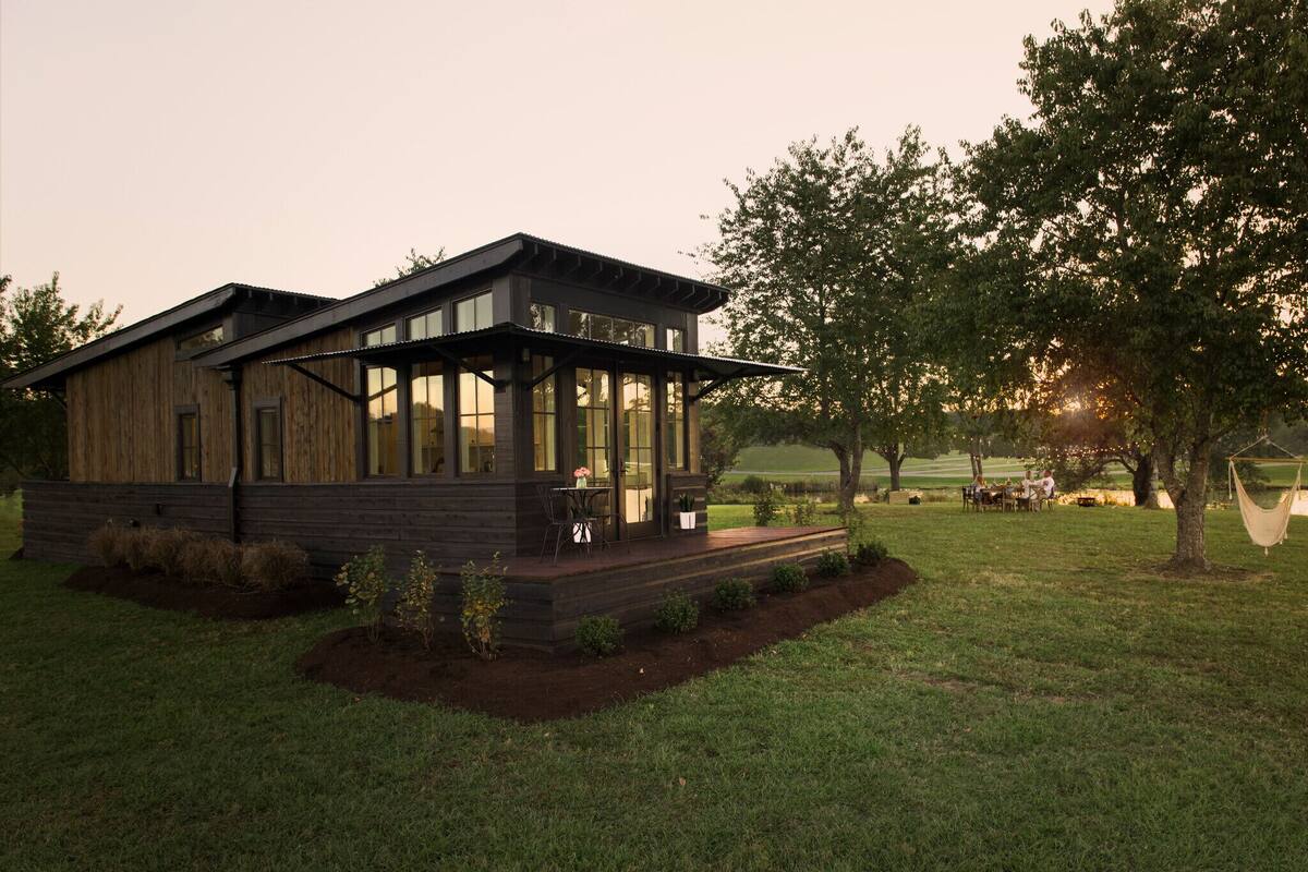 A modern wooden cabin with a glass front, a deck, and a view of a grassy field and trees.