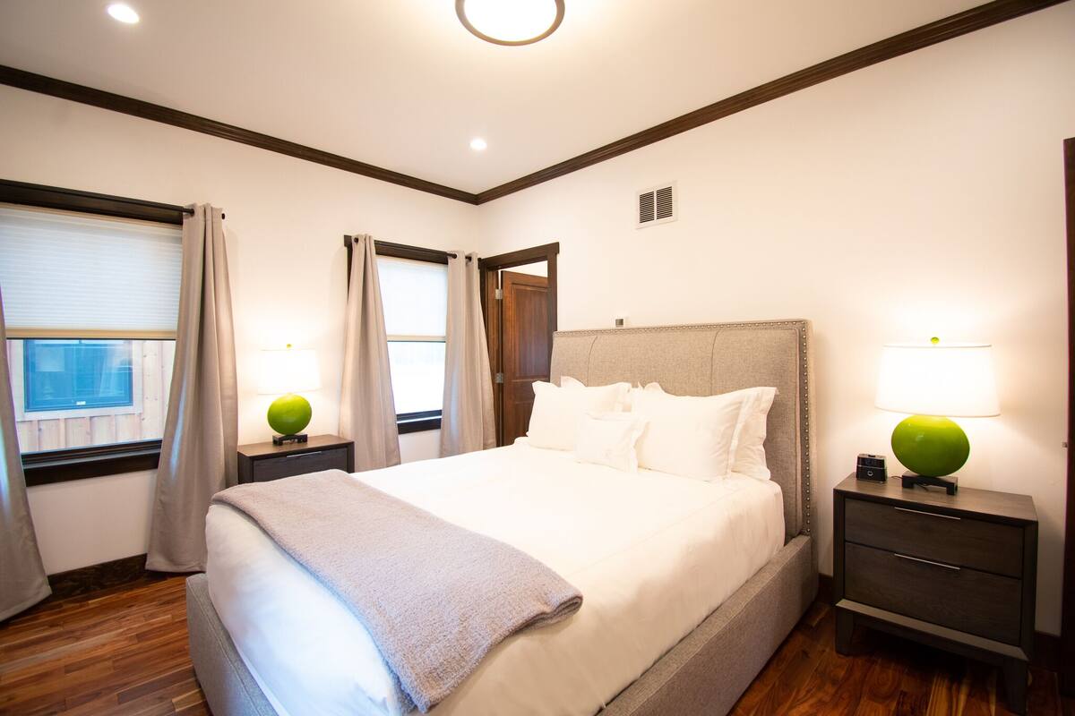 A hotel room with a large bed, two bedside tables with lamps, a window with curtains, and a wooden floor.