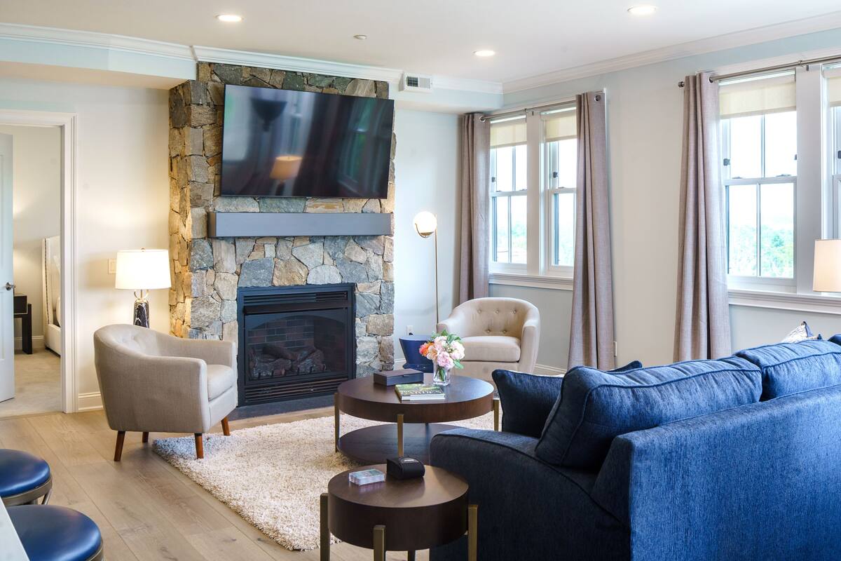 A living room with a stone fireplace, a flat-screen TV mounted above it, a beige armchair, a blue sofa, and a round coffee table with a vase of flowers.