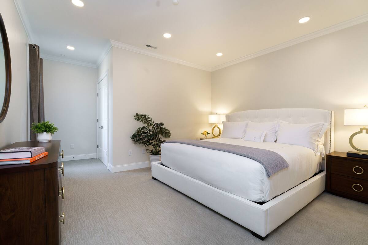A bedroom with a white bed, bedside tables, a nightstand, a potted plant, and a mirror on the wall.