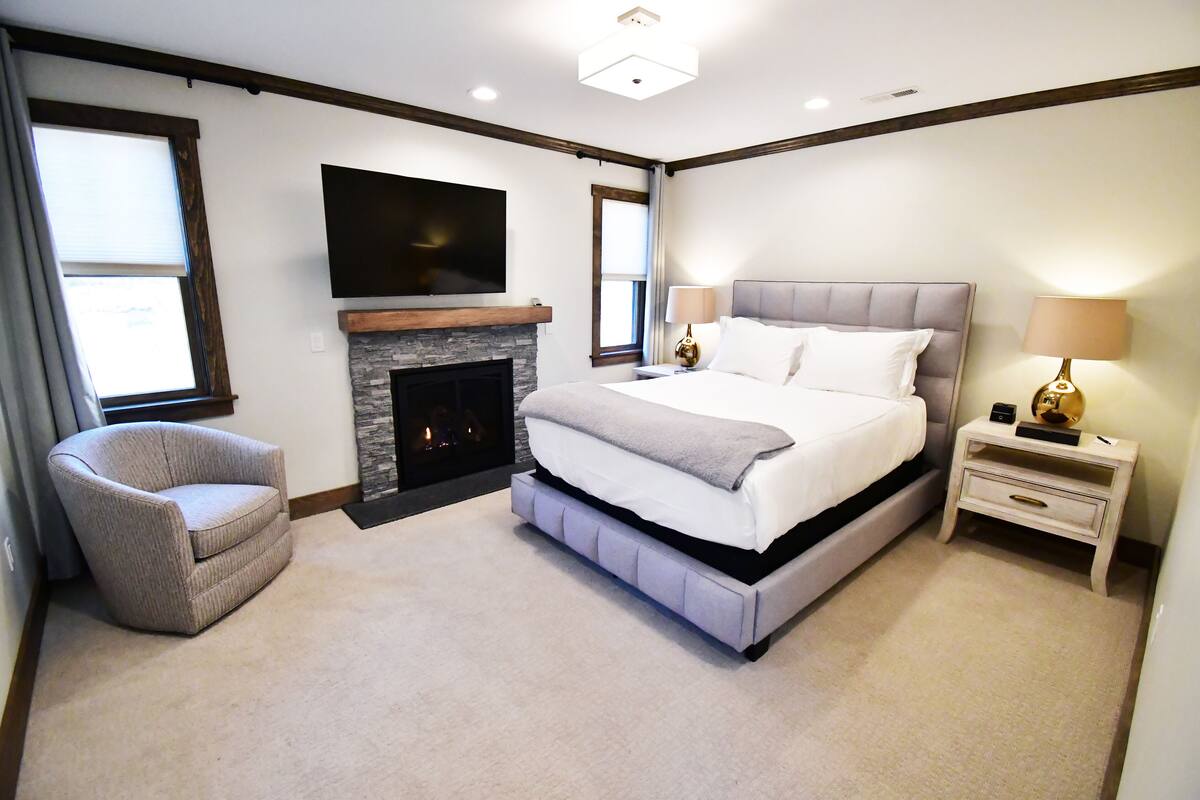 A bedroom with a large bed, a TV mounted on the wall, a fireplace, and a chair.