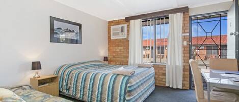 A hotel room with a bed, a bedside table with a lamp, a chair, a desk with a chair, a window with curtains, and a picture on the wall.
