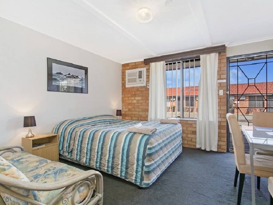 A hotel room with a bed, a bedside table with a lamp, a chair, a desk with a chair, a window with curtains, and a picture on the wall.