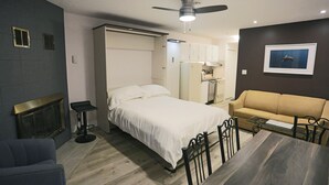 A bedroom with a bed, a sofa, a dining table, a ceiling fan, a fireplace, and a framed picture on the wall.