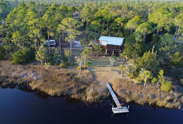 An aerial view of a cabin surrounded by dense forest, a dock extending into a body of water, and a clear path leading to the cabin.