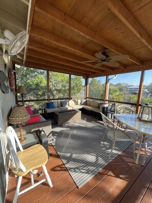 A covered outdoor seating area with wooden flooring, a sofa set, a dining table, and a ceiling fan.