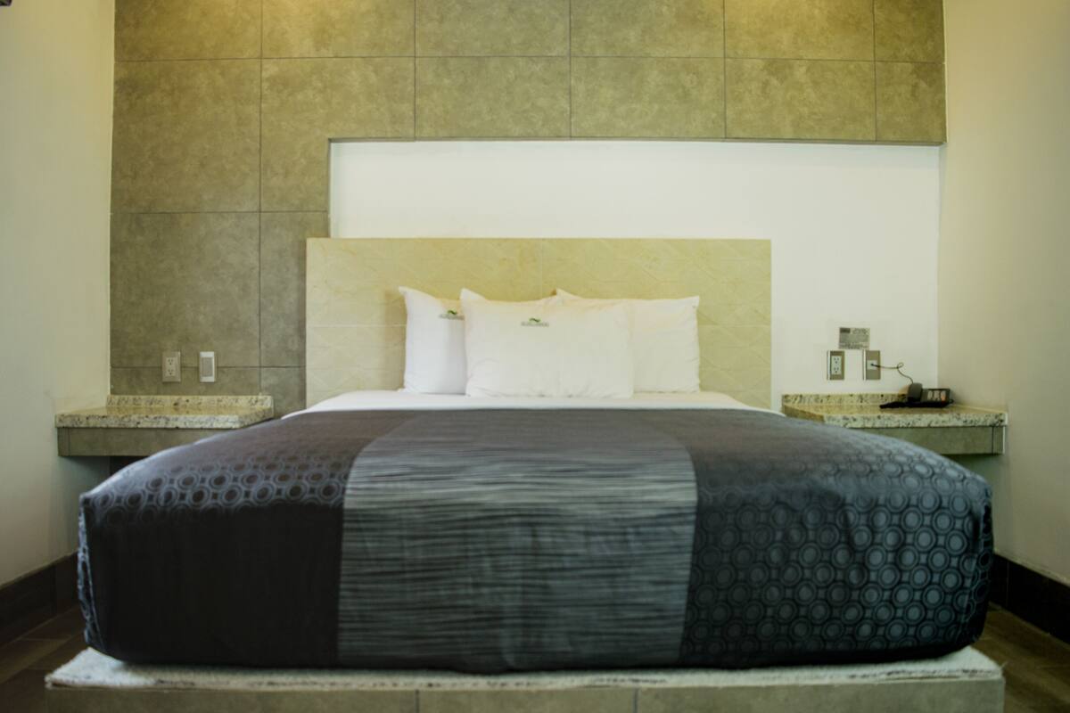 A hotel room with a large bed, a headboard, a nightstand, and a wall-mounted light.