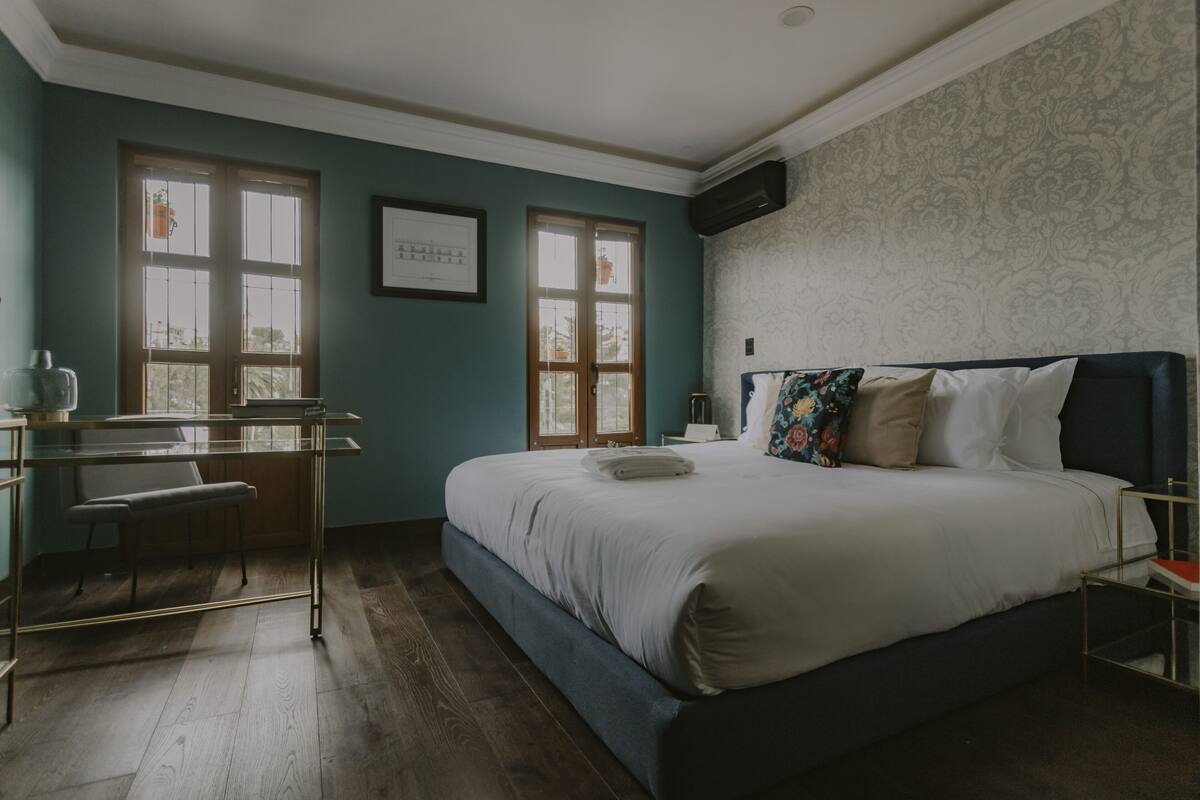 A bedroom with a bed, a desk, and a chair. There are two windows with curtains, a wall-mounted air conditioner, and a framed picture on the wall.