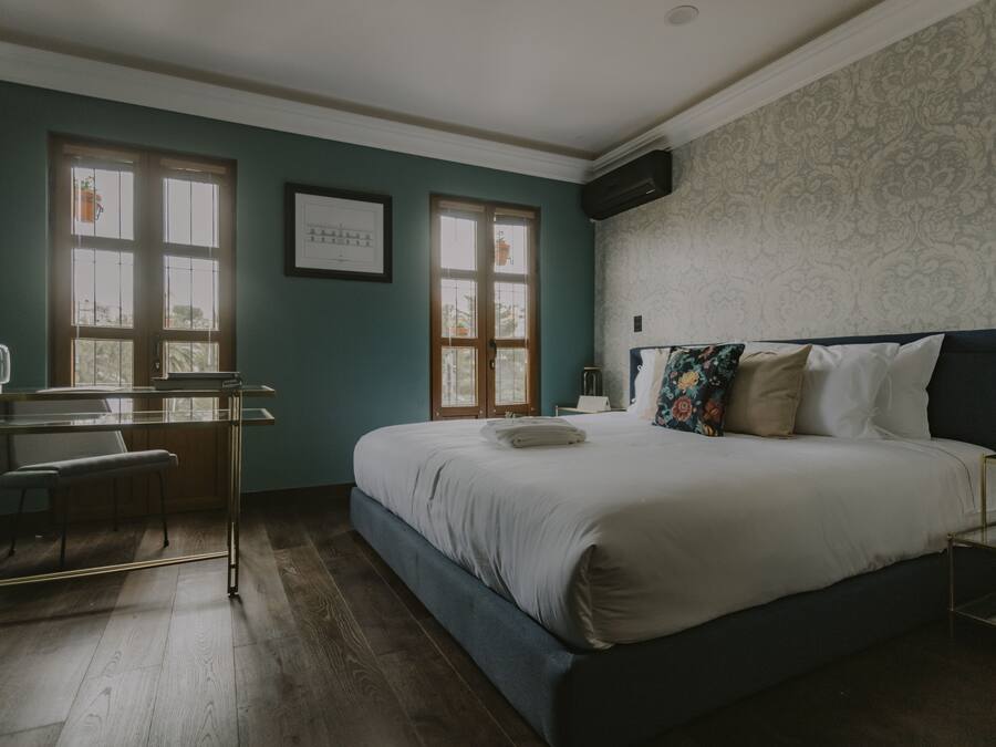 Ein Schlafzimmer mit einem Bett, einem Schreibtisch und einem Stuhl. Zwei Fenster mit Vorhängen, eine Wand-Klimaanlage und ein gerahmtes Bild sind zu sehen.