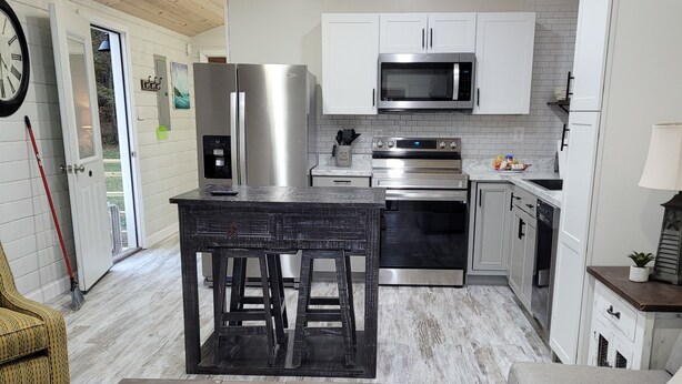 A compact kitchen with white cabinets, stainless steel appliances, and a dining area with a rustic table and stools. - Crossroads to Pickwick Cabin SMOOTH SAILING (Iuka)