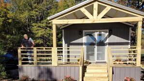 A wooden structure with a covered entrance, gravel ground, and a person standing on the porch. - Crossroads to Pickwick Cabin SMOOTH SAILING (Iuka)