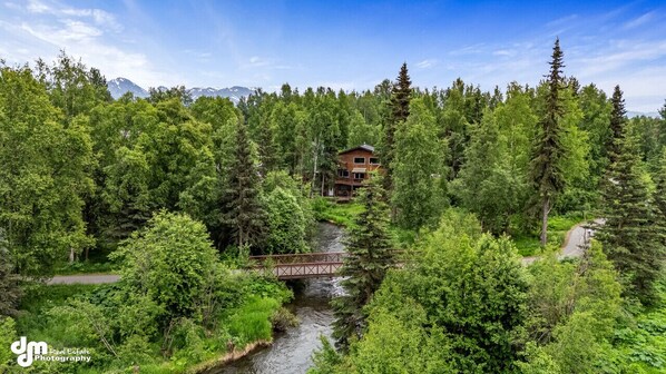 A two-story lodge surrounded by dense forest, a bridge crossing a stream, and mountains in the background.