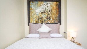 A neatly made bed with a patterned bedspread, a large framed painting of a horse above it, and a bedside lamp on a wooden nightstand. - Palmer House, Studio 2, Cozy Downtown Space, Sleeps 4! (Baker City)