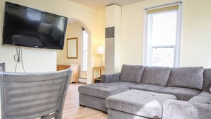 A modern living room with a sectional sofa, a flat-screen TV mounted on the wall, a wooden chair, and a small table with a lamp. - Palmer House, Studio 2, Cozy Downtown Space, Sleeps 4! (Baker City)