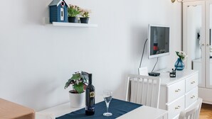 Un coin repas avec une table blanche, une nappe bleue et des chaises blanches avec des coussins bleus. Il y a une horloge au mur, une petite décoration représentant une maison et un téléviseur posé sur un meuble.