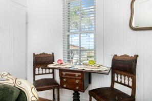 Une pièce avec un mur recouvert de panneaux blancs, une fenêtre avec des stores, une table en bois recouverte d’une nappe à motifs floraux, deux chaises en bois et un miroir accroché au mur.