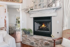 Una chimenea de piedra con fuego, un sofá blanco y un comedor al fondo.
