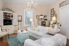 Una habitación con un sofá blanco, una mesita con una lámpara, un televisor antiguo y una araña.