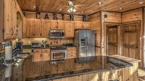 A cabin-style kitchen with wooden cabinets, a stainless steel refrigerator, a microwave, a stove, and a granite countertop.