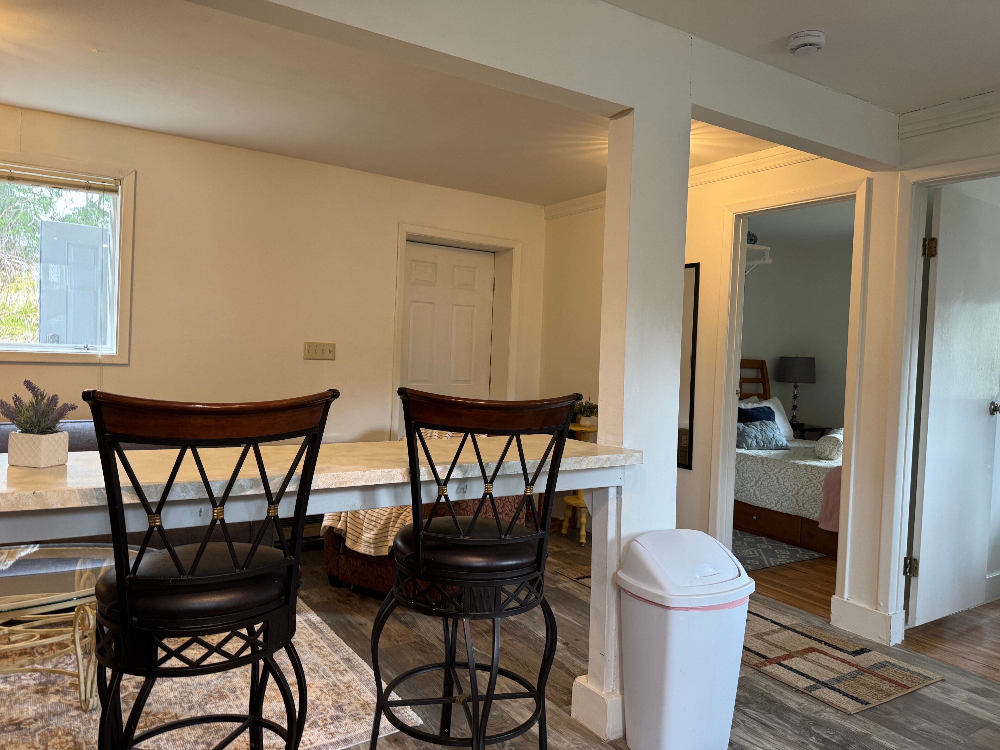 Una cocina con dos taburetes de bar, un bote de basura blanco y vista a un dormitorio.