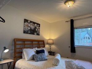 A bedroom with a wooden headboard, a bed with white and patterned bedding, a nightstand with a lamp, a painting of flowers, and a window with a view of a house. - Walk Almost Anywhere at Alyeska Resort when you stay at Chair Four Por Favor! (Girdwood)