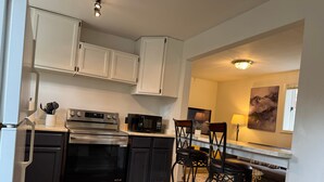 A compact kitchen with a stove, microwave, and white cabinets. There are two bar stools and a trash can. - Walk Almost Anywhere at Alyeska Resort when you stay at Chair Four Por Favor! (Girdwood)