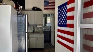 A kitchen with white cabinets, a black refrigerator, and an American flag on the wall. - Biker's Bungalow - Near Mendenhall Glacier and Auke Bay Offering DISCOUNT ON TOURS! (Juneau)
