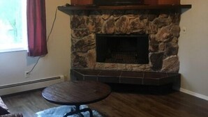 A living room with a stone fireplace, a flat-screen TV mounted above it, a round wooden table, and a fur rug. - Sand Lake Jewel - Near Airport, Shopping and the Best Sushi in Town! (Anchorage)