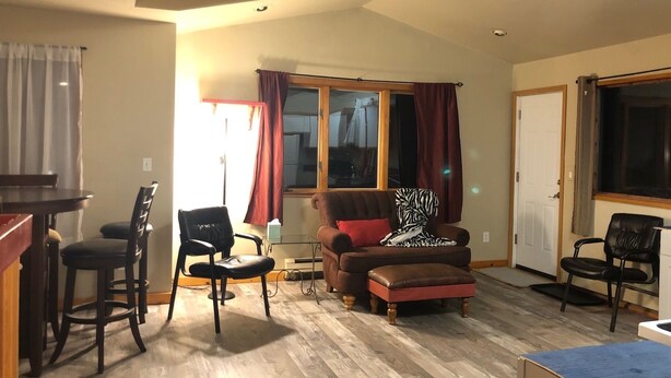 Un salon comprenant un fauteuil marron, un petit pouf, une table basse en bois et un coin repas avec une table et des chaises.