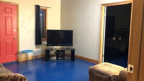 Une pièce avec un téléviseur à écran plat posé sur un meuble, une porte rouge, une petite chaise bleue et un pouf à motifs.