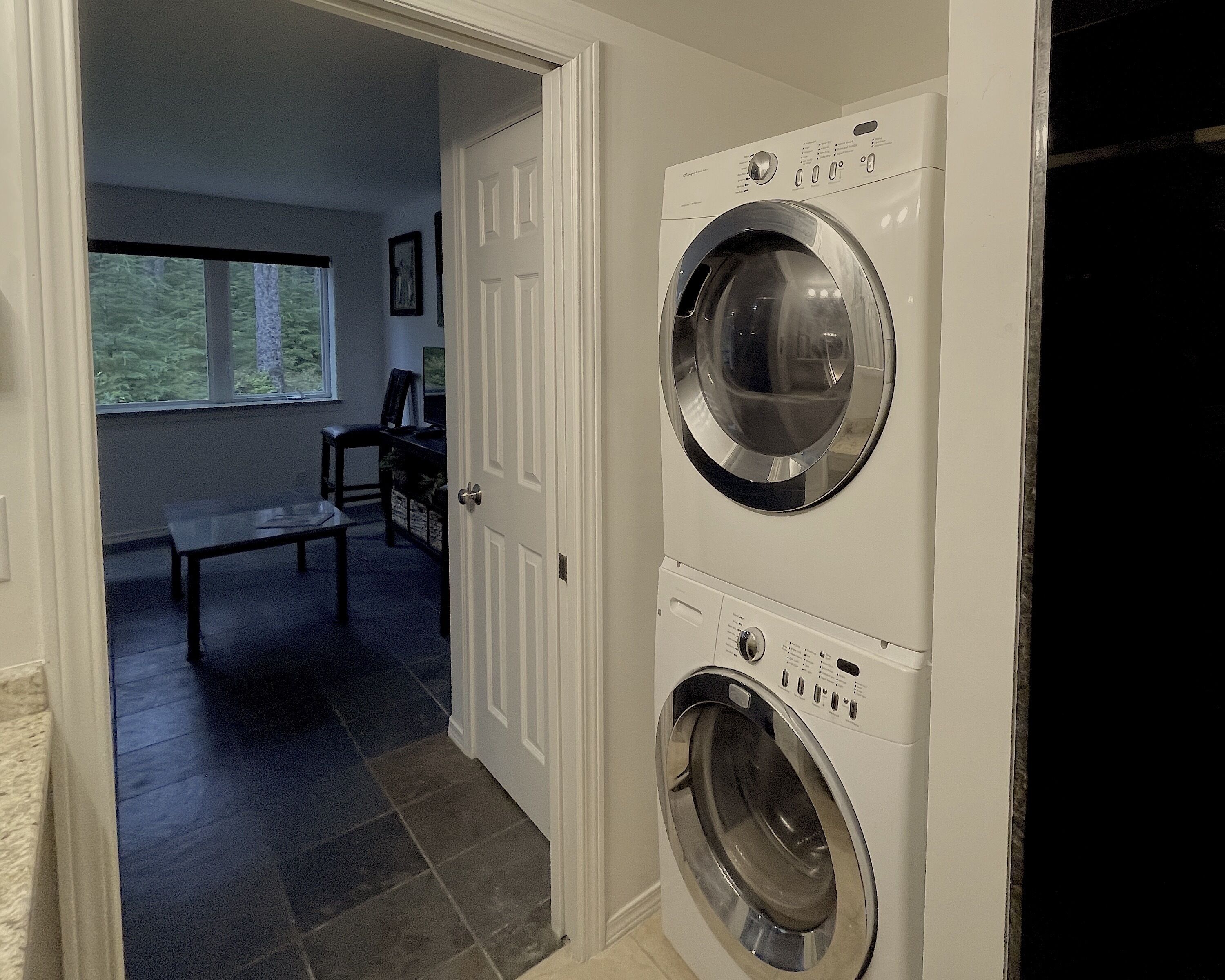 A laundry room with a front-loading washer and dryer, a bench, and a window with a view of trees.