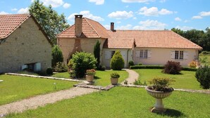 Ein Steinhaus mit rotem Ziegeldach, umgeben von einem gepflegten Garten mit Wegen und Kübelpflanzen.