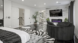 Un dormitorio moderno con una cama, un tapete estampado de cebra, un sillón gris y un televisor montado en la pared.
