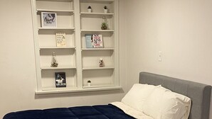 A bedroom with a bed, a white headboard, a window with shelves, and a wooden floor.