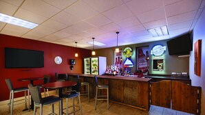 Eine Barbereich mit mehreren Tischen und Stühlen, ein mit Getränken gefüllter Kühlschrank und ein an der Wand befestigter Fernseher.