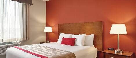 A hotel room with a large bed, two bedside tables with lamps, a window with curtains, and a red accent wall.
