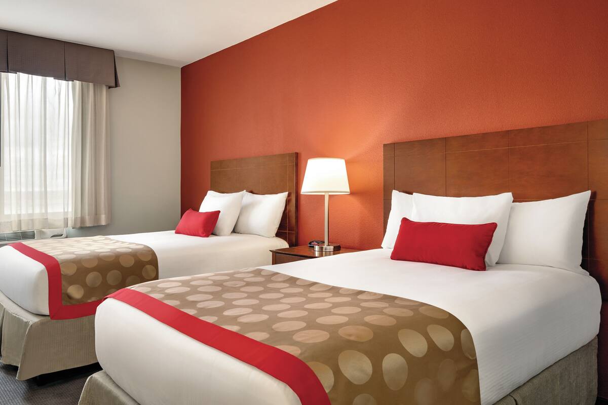 Habitación de hotel con dos camas, cada una con un cojín rojo, una mesita de noche con lámpara y una ventana con cortinas.