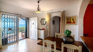 A dining area with a wooden table, white chairs, a large vase, and a view of the sea through a glass door. - House of the Rising Sun - Luxury villa with private pool and stunning sea views (Sayalonga)