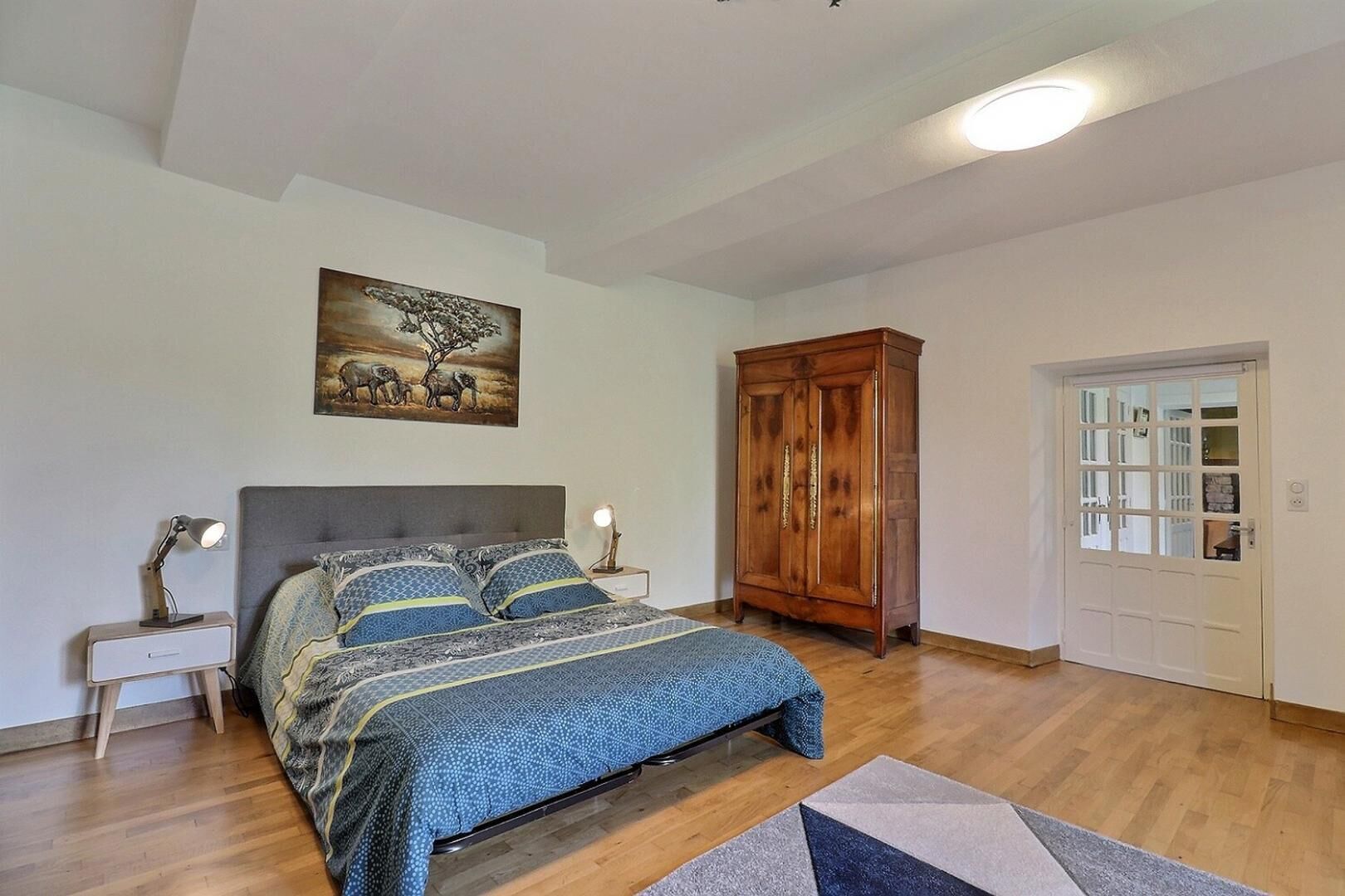 Een slaapkamer met een bed, nachtkastjes, een houten garderobe, een deur en een schilderij aan de muur.