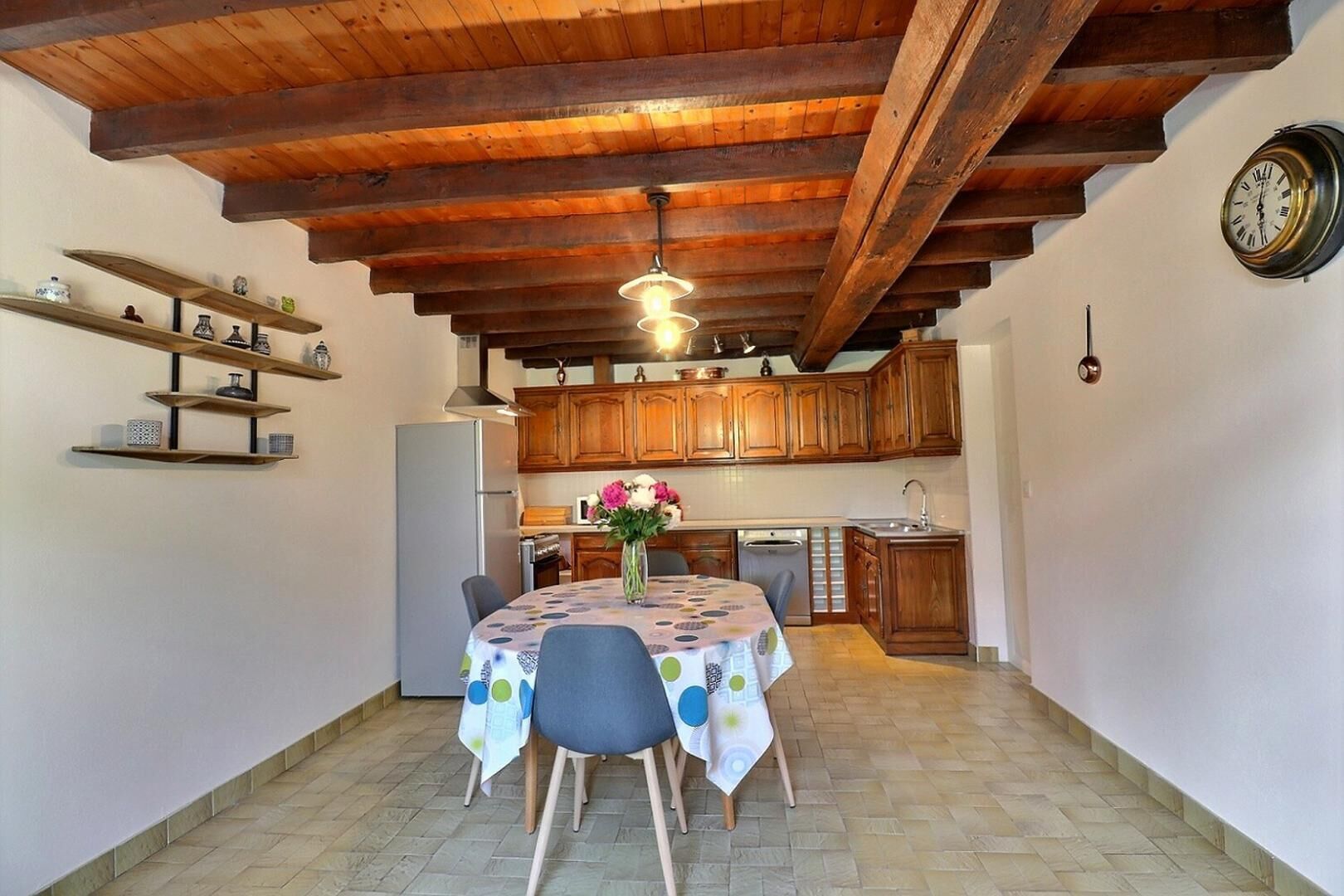 Une cuisine compacte avec des meubles en bois, une table à manger avec des chaises, un réfrigérateur et une horloge au mur.