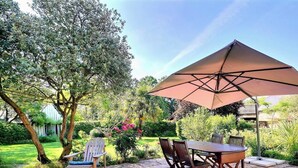 Une terrasse avec une table et des chaises, un grand parasol, et un jardin avec différentes plantes et arbres.