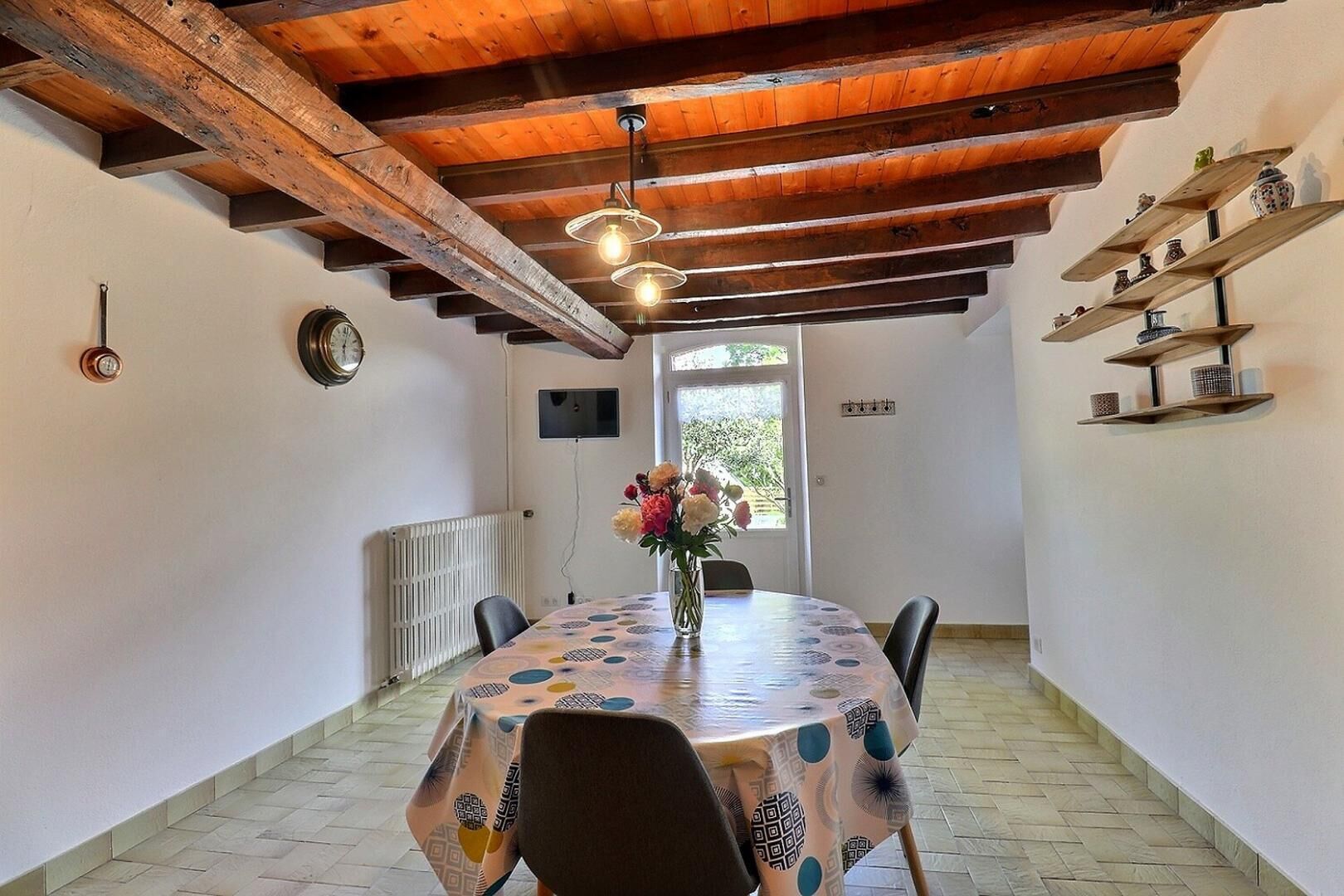 Ein Essbereich mit Holzboden, einem runden Tisch mit gemustertem Tischläufer, Stühlen, einer Vase mit Blumen und Regalen mit Dekoration.