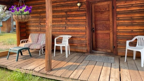 Eine Holzhütte mit einer Veranda, zwei weißen Plastikstühlen, einer Bank und einem hängenden Blumenkorb.