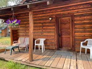 Een houten chalet met een veranda, twee witte plastic stoelen, een bankje en een hangmand met bloemen.