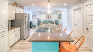 A kitchen with white cabinets, a stainless steel refrigerator, a granite countertop island, and orange chairs. - Stay 7+ Nights for 10% Off: Isla Breeze, Your Easeful Escape (South Padre Island)